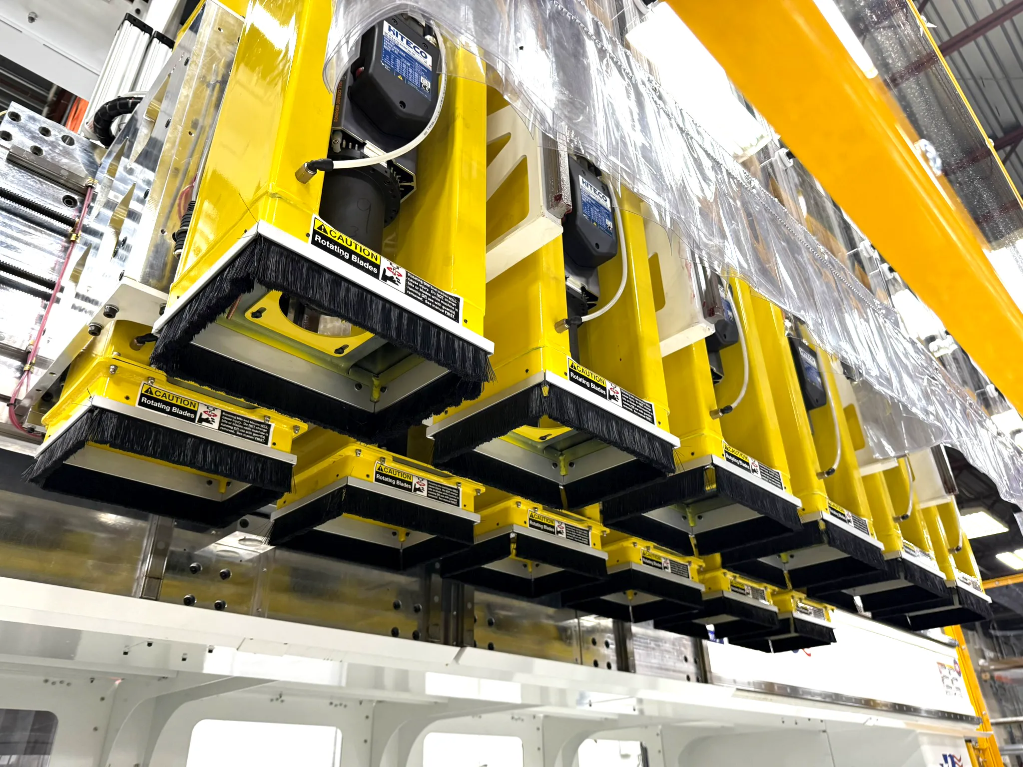 Close-up underside view of multiple yellow CNC routing heads, each equipped with black bristle dust skirts and "Caution – Rotating Blades" warning labels. The units are branded with "HITECO" motor labels and are mounted in a grid-like array.