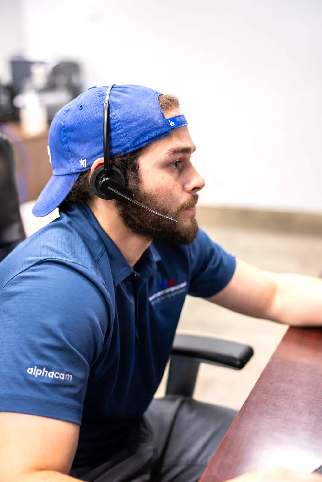 A young male technician wearing a blue "alphacam" polo shirt, a backwards LA Dodgers cap, and a wireless headset, focused on a screen while providing technical support or remote assistance.